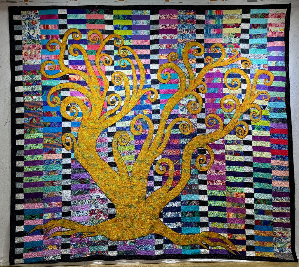 A vibrant quilt featuring a stylized tree design in shades of yellow against a colorful striped background made of various fabrics.