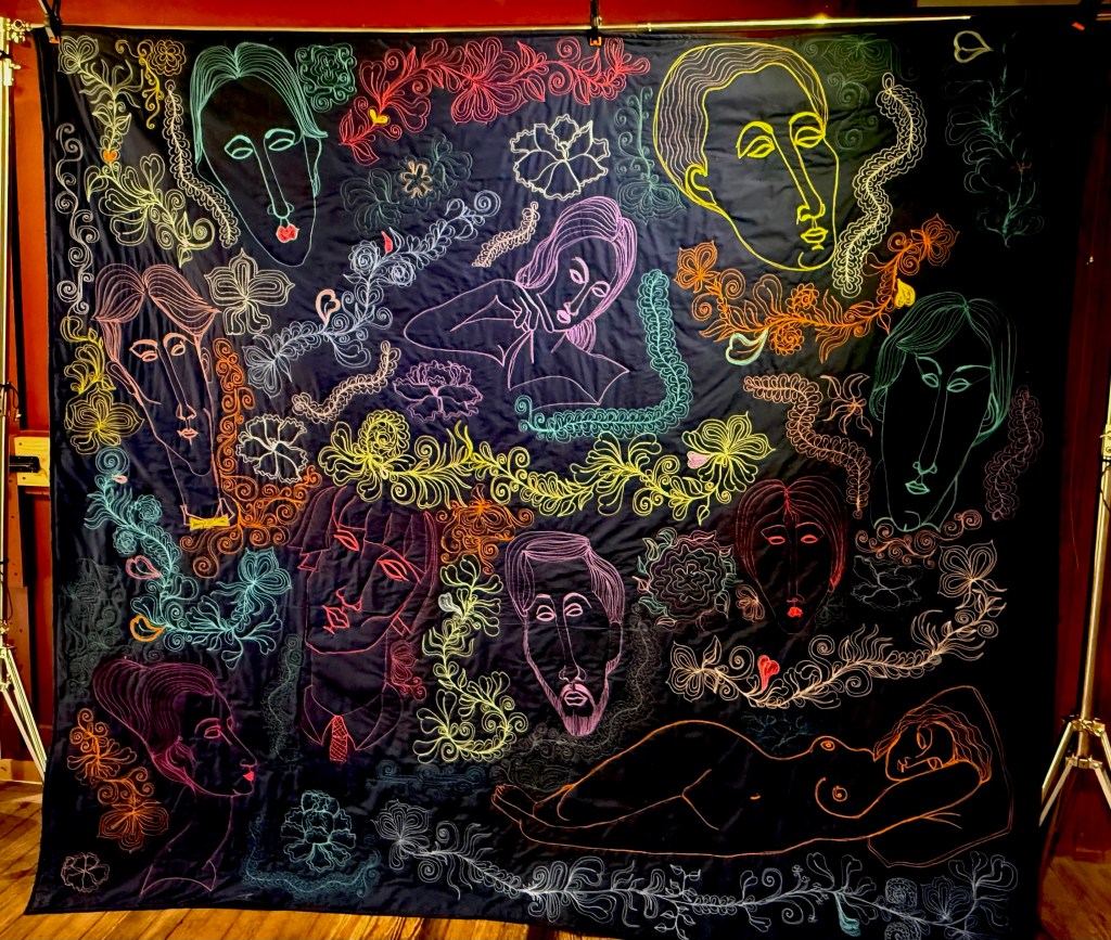 A colorful quilt titled 'Family Portrait' featuring outlined faces and floral designs, showcasing various expressions and intricate patterns on a dark background.