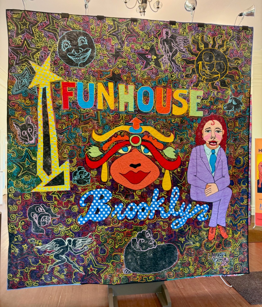 A vibrant quilt titled 'Funhouse' featuring colorful designs, including a large face, stars, and playful motifs, displayed in an exhibition space.