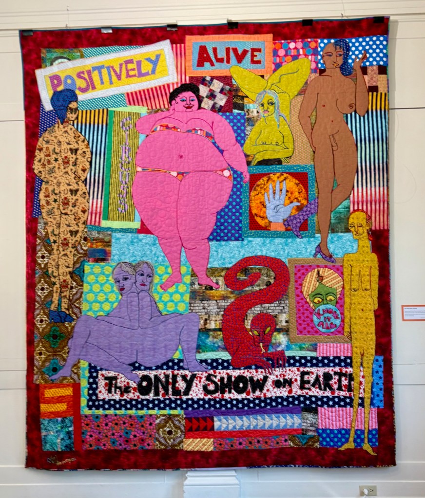 A colorful quilt titled 'Positively Alive' featuring various stylized human figures in vibrant fabrics, accompanied by playful text and motifs.