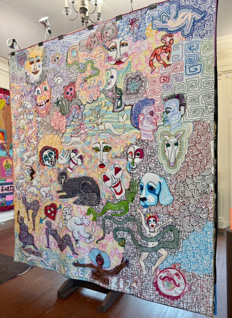 A colorful quilt featuring abstract faces, playful figures, and various whimsical designs, displayed on an easel in a well-lit room.