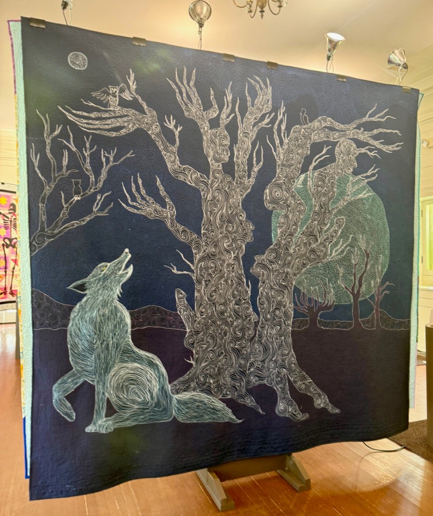 A quilt featuring a howling wolf in front of a stylized tree with intricate swirling patterns and a rising moon, showcased in an indoor exhibit.