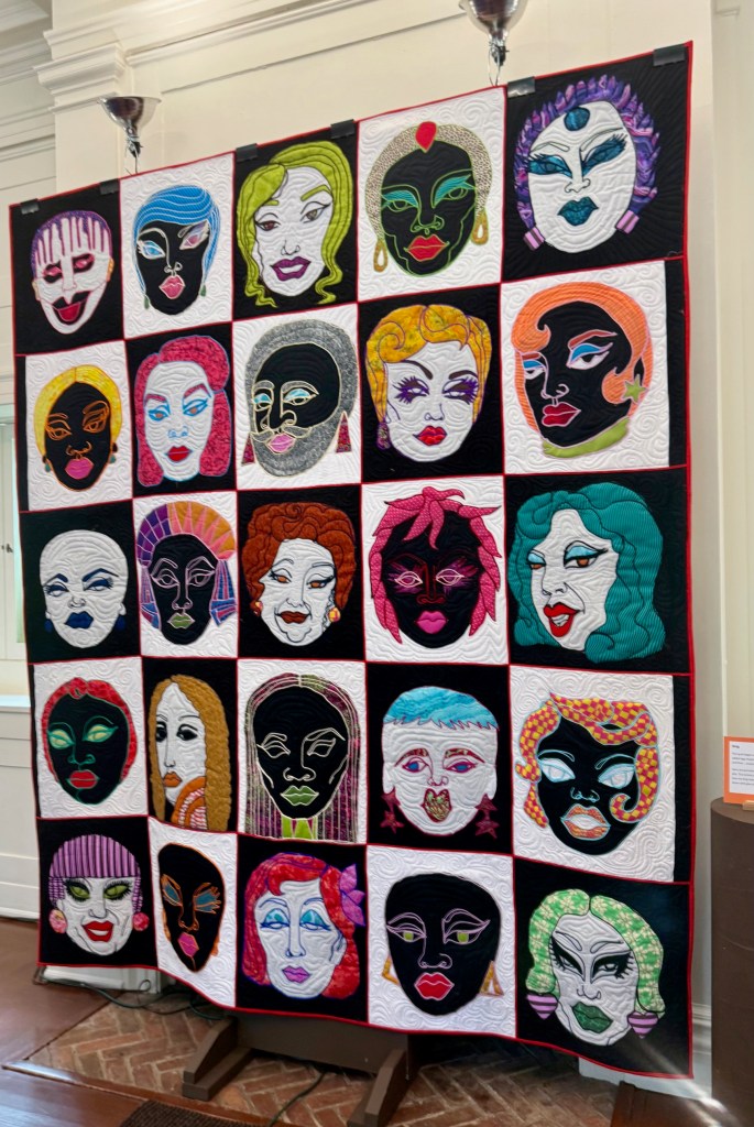 A quilt display featuring a grid of colorful, stylized faces, each with unique hairstyles and makeup, on a black background.