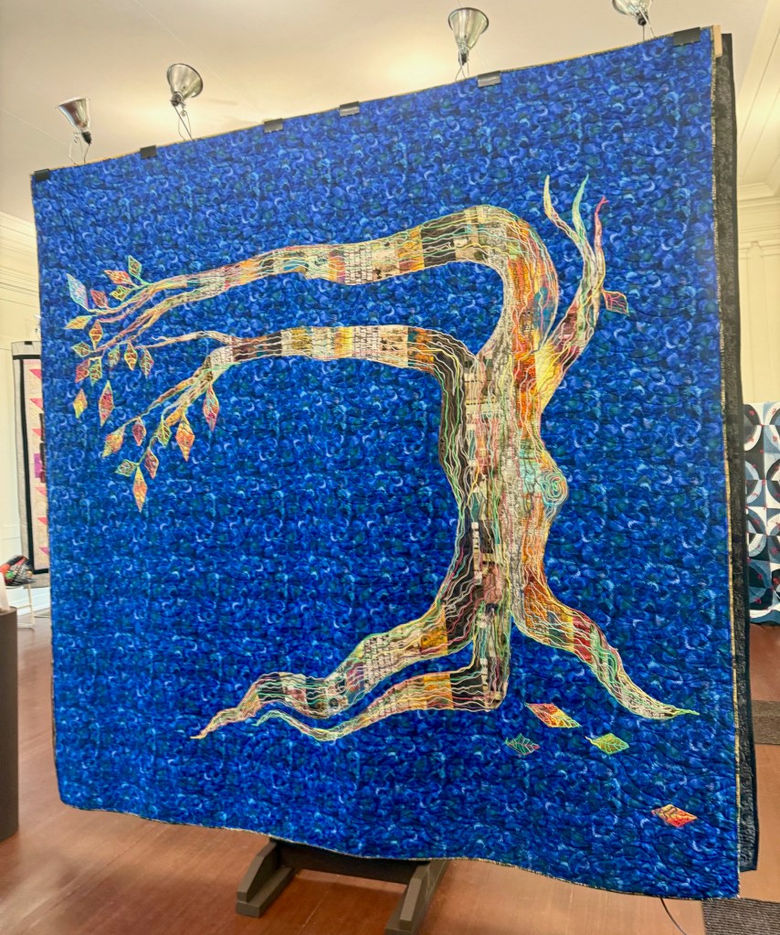 A colorful quilt titled 'Walking Tree,' featuring a stylized tree design with branches extending upwards and vibrant leaves against a blue background.