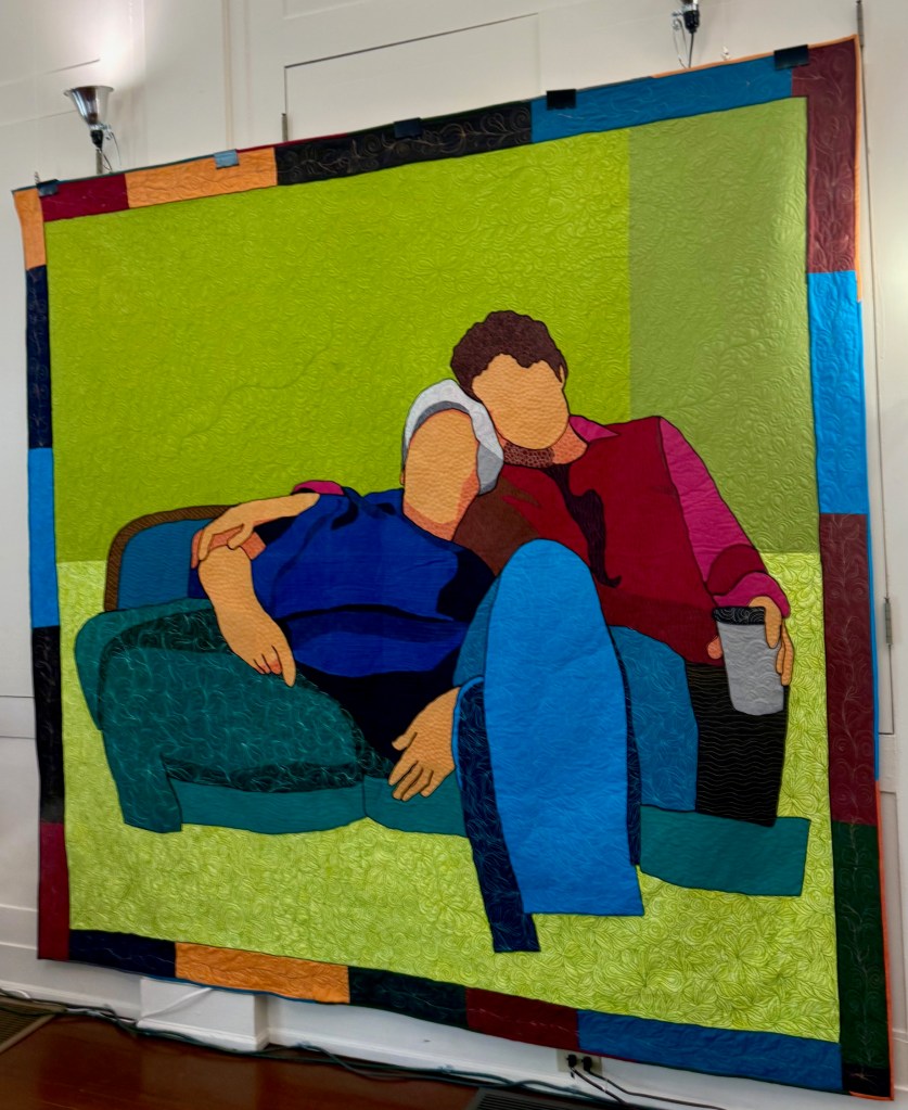 A colorful quilt featuring two figures sitting closely on a couch, with vibrant fabrics depicting their clothing and the couch, against a bright green background.