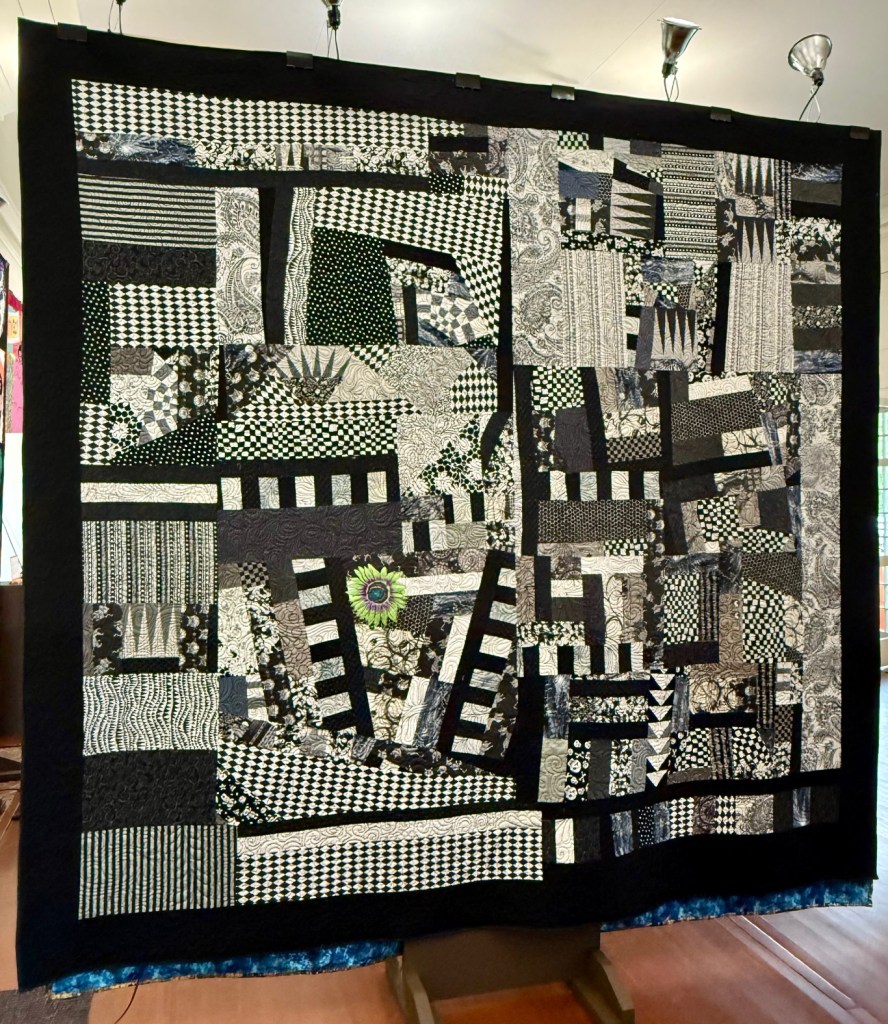 A striking black and white quilt featuring various patterns and textures, including florals and geometrics, showcased in a gallery setting.