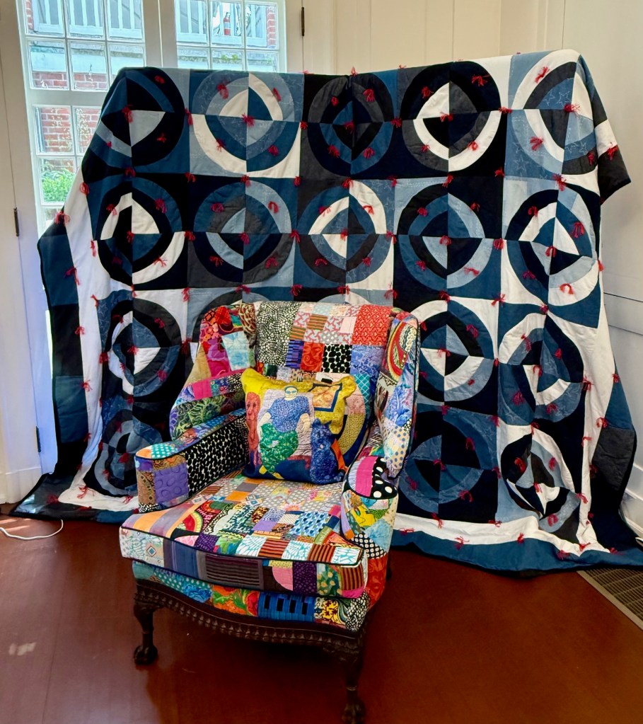 A brightly colored, patchwork upholstered chair in the foreground with a quilt featuring a pattern of circles in blue, white, and dark blue hues hanging in the background.