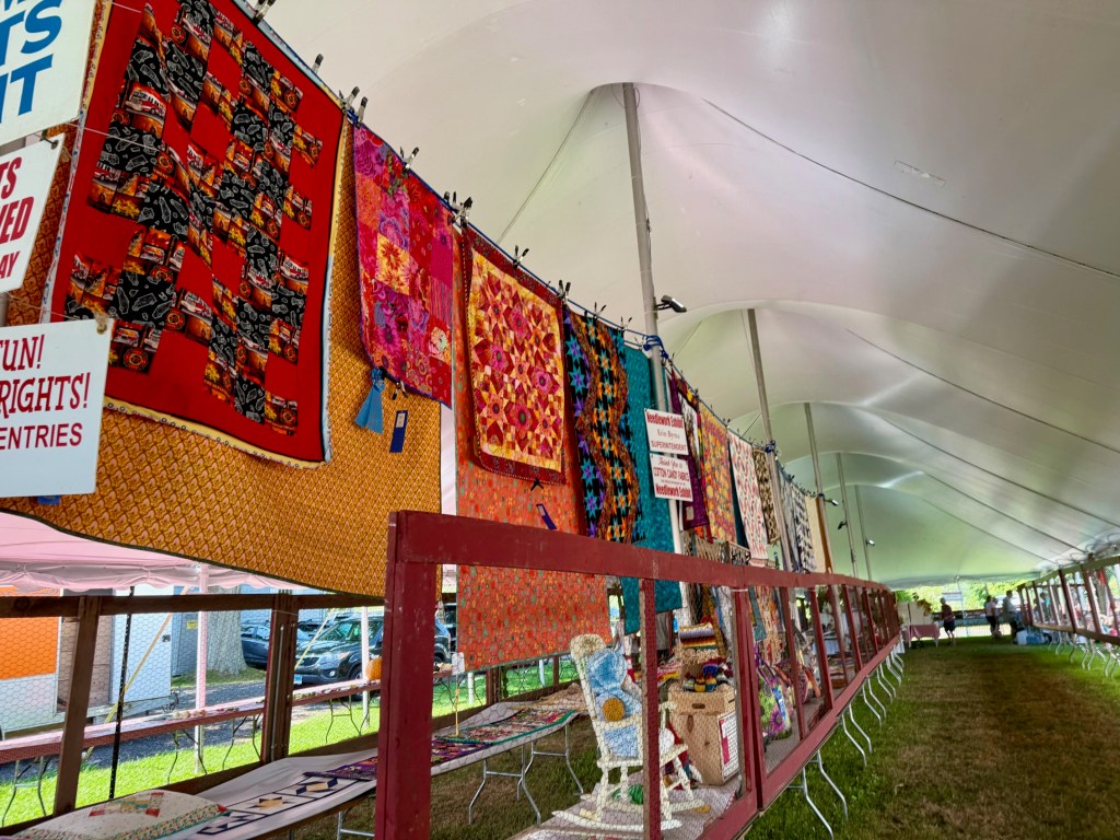 Colorful quilts on display under a tent at a fair, showcasing various designs and patterns, with a sign indicating the Needle Arts exhibit.